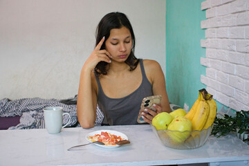 Young Indian woman at a table in a gray tank top, looking at her smartphone, breakfast food and fruit on the table. Perfect for morning tech use, wellness, or digital lifestyle visuals.