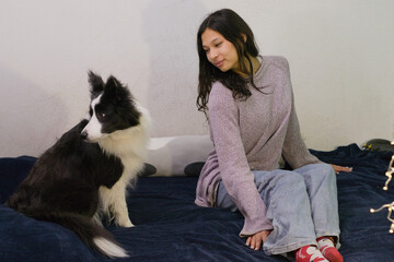 Woman sitting on a bed, wearing comfortable clothes, looking at her black and white dog who is also on the bed. Ideal for calm pet moments or home relaxation themes.
