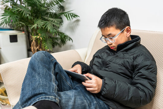 A young boy is seated on a sofa, engrossed in using a digital tablet for leisure in an indoor environment. - Powered by Adobe