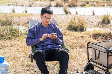 Obraz premium A man is sitting outdoors on a chair, using his mobile phone in a natural setting.