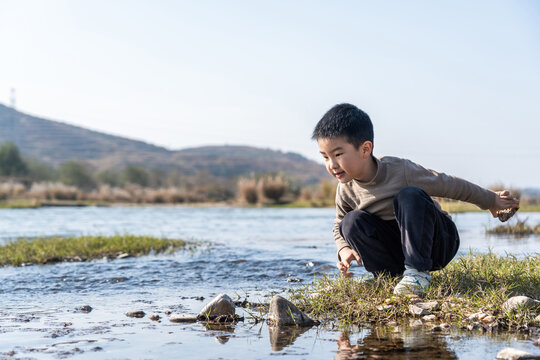 A young boy is crouching by the river, exploring the water in a natural outdoor environment.