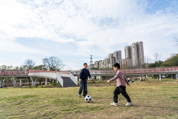 Two individuals are engaged in a soccer game on a grassy field with buildings in the background under a partly cloudy sky.