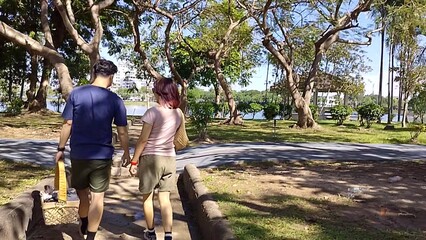 Back view of a couple walking on a sunny park path holding a basket and bag. They enjoy a relaxing stroll near green trees and a lake during summer vacation.