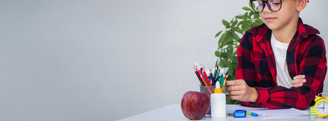 Smart young boy in glasses preparing for school, choosing pencils from the organizer on his desk.