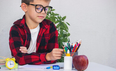 Smart young boy in glasses preparing for school, choosing pencils from the organizer on his desk.