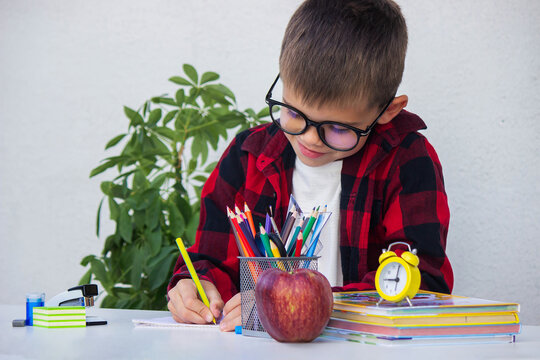 Child writing or drawing at a desk surrounded by school supplies, books, an apple,and an alarm clock