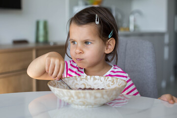 young girl in a striped shirt eating cereal at a kitchen table. Child enjoying breakfast in a bright modern home interior, healthy morning routine concept. close up portrait