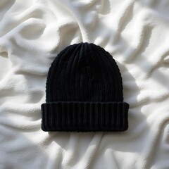 Black knitted beanie hat with a ribbed cuff, resting on a soft white textured fabric surface with subtle shadows. black beanie