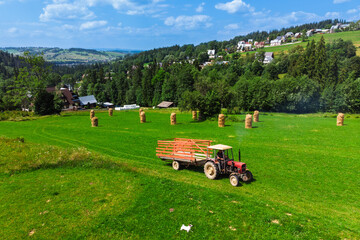 Tractor working in a field during haymaking in Podhale, Poland. Summer rural landscape featuring agricultural work, green hills, and traditional houses.