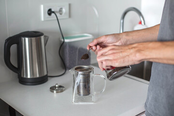 A person preparing tea in a modern kitchen, adding loose leaf tea into a glass teapot with a metal infuser next to an electric kettle. Home beverage preparation concept.