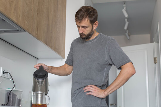 A man in a modern kitchen pouring hot water from an electric kettle into a glass teapot, preparing a hot beverage. Home brewing and daily routine concept. morning routine