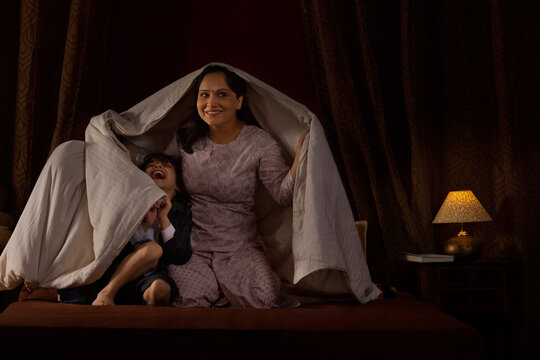 Happy mother playing with her two little kids with blanket on bed