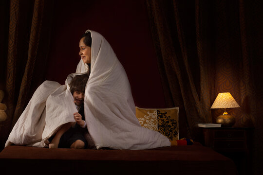Happy mother playing with her two little kids with blanket on bed