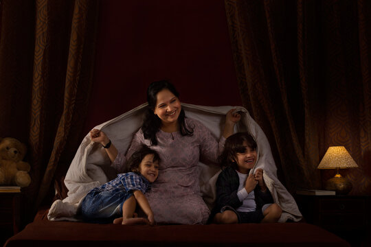 Happy mother playing with her two little kids with blanket on bed