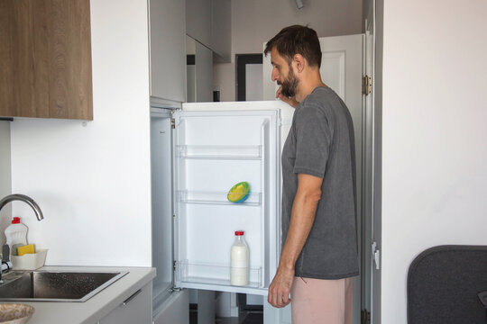 man standing in a modern kitchen looking into an open refrigerator with minimal food inside. Concept of empty fridge, grocery shortage, meal planning, and domestic lifestyle.
