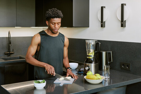 Young adult Black man preparing breakfast by slicing onion on cutting board in modern kitchen, standing near blender and fresh fruit, focused on morning routine