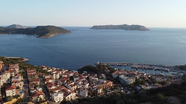 Aerial drone shot showing red tiled rooftops on hillside Kalekoy overlooking Kekova waters with island views, calm blue sea, scattered homes, warm daylight, and peaceful coastal atmosphere