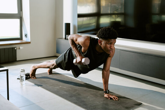 Young adult Black man exercising on yoga mat performing one arm push up with dumbbell in modern living room during morning workout routine, focused expression, looking forward
