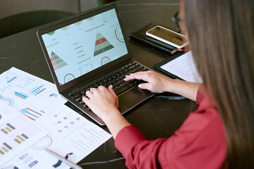 Caucasian young adult woman working on laptop analyzing business charts and graphs in morning, hands typing on keyboard, financial documents and smartphone on desk, focusing on data visualization