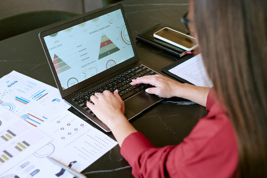 Caucasian young adult woman working on laptop analyzing business charts and graphs in morning, hands typing on keyboard, financial documents and smartphone on desk, focusing on data visualization