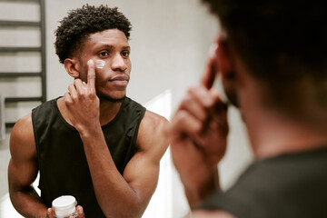 Young adult Black man applying facial cream to cheek while standing in front of mirror, holding...