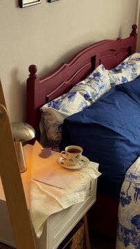 Cozy close-up of a bedside table with a cup of hot tea, a book, and a glowing lamp, set next to a bed with dark blue and white patterned Toile de Jouy bedding and a burgundy headboard, depicting relax