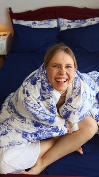 A cheerful young woman happily wrapped in a white and blue Toile de Jouy patterned duvet, sitting comfortably in fresh, dark blue bedding, conveying coziness, domestic relaxation, and a joyful morning