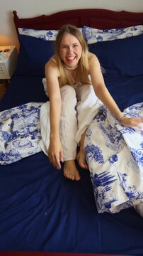 A cheerful, playful woman in white pajamas jumping onto a bed with dark blue and white Toile de Jouy patterned bedding, raising her feet up in the air and laughing, conveying fun, relaxation, and happ