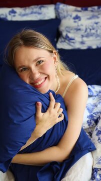 Close-up of a beautiful young blonde woman smiling happily while hugging a soft blue pillow in a bed with dark blue and white Toile de Jouy patterned bedding, promoting comfort and relaxation