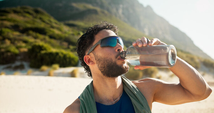 Fitness, break and man with drinking water by beach, hydration and sunglasses for summer training. Running pause, beverage and athlete with bottle for workout recovery, rest and outdoor with liquid