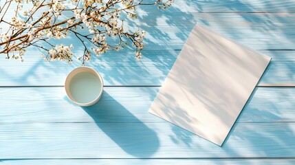 A flat lay composition featuring a blank white paper, a cup of liquid, and a branch of white cherry blossoms on a light blue wooden table. Sunlight casts dapple