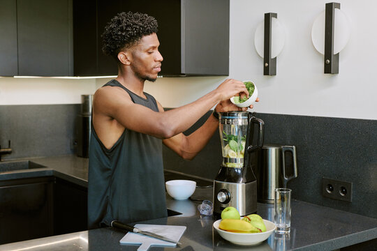 Young adult Black man preparing healthy smoothie in modern kitchen, placing fresh spinach and fruit into blender, standing at countertop with bowl, glass, and assorted fruit nearby