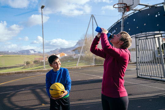 Caucasian mother drinking water from a bottle while son holding a basketball looks at her on an outdoor court - Powered by Adobe