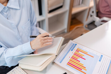 A young Asian businesswoman is sitting at her desk in a modern office, reviewing printed documents while working on a computer, showing focus, dedication, and professional attitude.