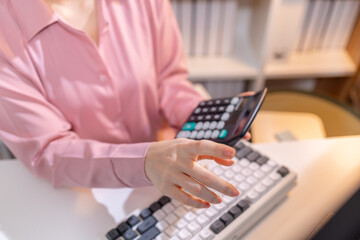 A young Asian businesswoman is sitting at her desk in a modern office, reviewing printed documents while working on a computer, showing focus, dedication, and professional attitude.