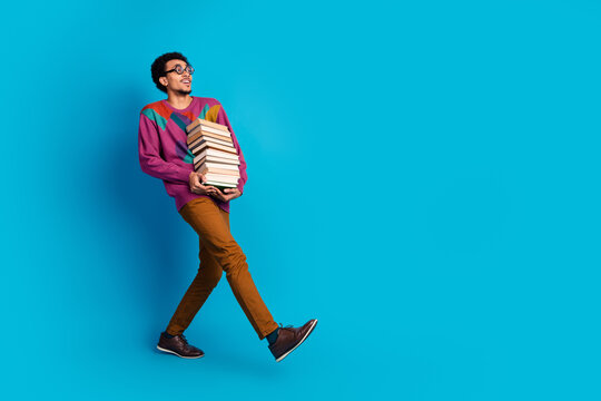 Young man carrying a stack of books while walking on blue background showcasing leisure learning and style