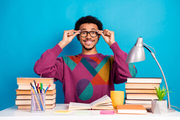 Cheerful young student adjusting glasses at study desk surrounded by books and stationery against a vibrant blue background