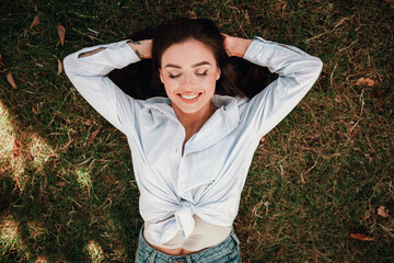 Playful woman lies on grass in a casual tied shirt and jeans enjoying a sunny day in the park chic...
