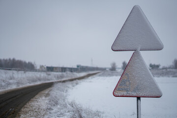 Snow covered warning road sign beside rural road in winter landscape with open fields background © Adam