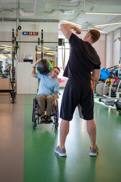 Disabled man lean to side to limit with medicine rubber ball, raise unilaterally, hold against forehead for cervical activation. Coach model correct posture breathing in workout, flexion conditioning