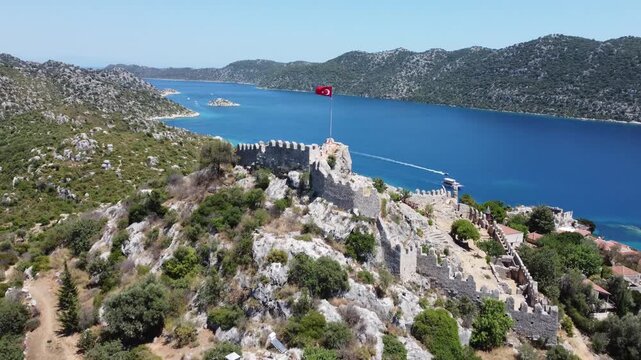Aerial drone shot showing crenellated fortress and red tiled village overlooking blue bay at Kalekoy in Antalya with rugged hills clear water warm daylight and calm coastal atmosphere