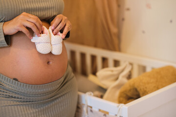 Pregnant woman holding baby shoes near crib at home. unrecognizable person