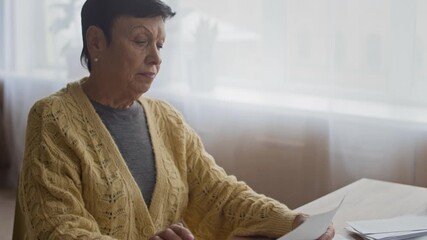 Tilt up of focused older Caucasian woman sitting at home with paperwork and calculator while checking bills and organizing monthly household finances - Powered by Adobe
