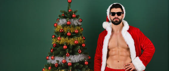 A stylish, muscular man in an open Santa suit and sunglasses stands confidently beside a richly decorated Christmas tree against a plain green background, creating a bold and playful holiday vibe.