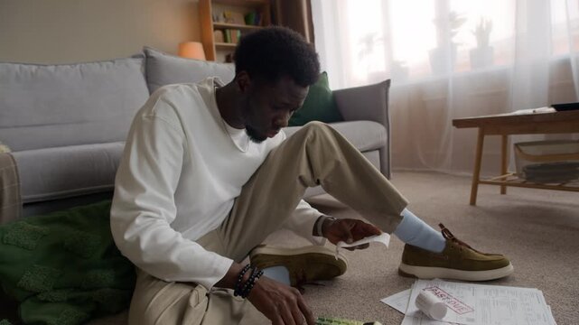 Young adult Black man sitting on floor at home reviewing receipts and overdue bills while calculating expenses with calculator in messy domestic setting