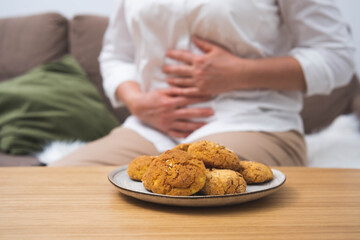 A plate of cookies in the foreground with a woman holding her stomach in discomfort in the background. Concept of overeating, food intolerance, stomach pain, or digestive issues.