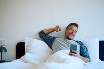 Smiling middle-aged man lying in bed after waking up, using smartphone and stretching, starting day relaxed