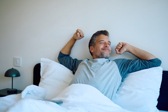 Man waking up in bed, stretching arms and smiling with closed eyes, feeling refreshed and happy. Enjoying a relaxing morning