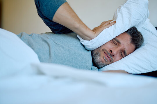 Male adult trying to block out loud noise with a pillow, experiencing sleep problems and stress in the morning