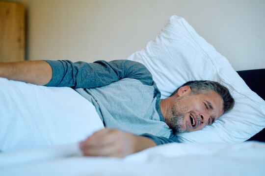 Man sleeping on his back on a bed, mouth open and facial expression showing a loud snore, indicating restless sleep, sleep issues, or a sleep disorder - Powered by Adobe
